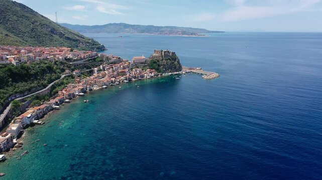 Slow aerial pan left along the coast of Scilla in Calabria showing a fortress on a promontory blue water rocks road and clear sky.