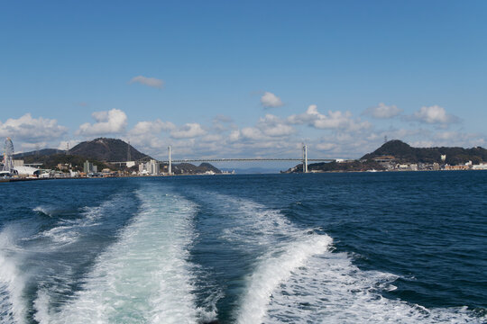 Panoramic view of Kanmon Bridge over the blue sea with white boat wake in the foreground under a clear sky with white clouds.