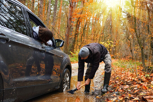 Man digging mud to free stuck car on forest road while another person watches from inside, autumn scenery with sunlight and fallen leaves