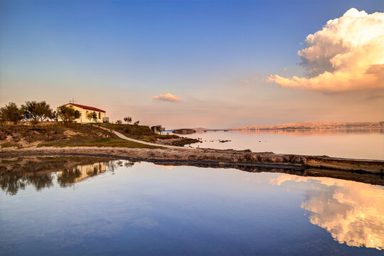 Lemnos Nea Koutali Saint Nicolas islet, Chapel standing on shoreline with bright clouds reflecting in tranquil water at sunset, creating a serene landscape