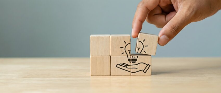 Hand completes a puzzle, carefully fitting the final wooden block to form a lightbulb cradled by an open palm, symbolizing the creation and nurturing of new ideas.