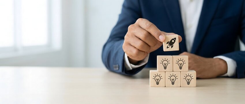 Hand places a wooden cube with a rocket icon on top of a pyramid of lightbulb cubes, signifying the launch of a new innovative idea.