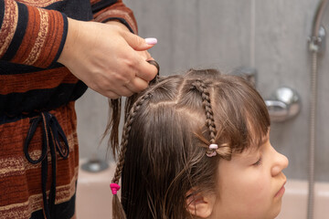 Naklejka premium Woman creates tidy hairstyle for child by braiding hair after washing at home.