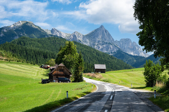 A winding road leads past traditional wooden houses and green meadows toward the sharp, towering peaks of the Austrian Alps under a blue sky in Austria.