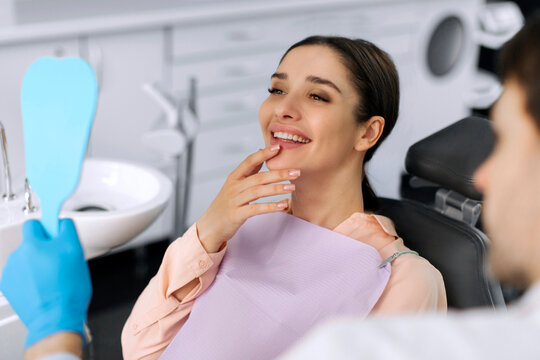Satisfied female patient sitting in dentist chair smiles after dental treatment while looking at her reflection in hand mirror