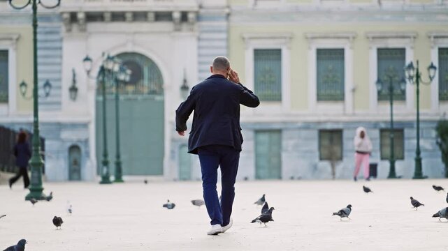 Businessman talk on phone walking on Kotzia square near historic building with pigeons in Athens, Greece