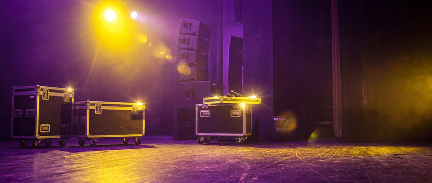 Professional flight cases and line array speakers on an empty stage during setup. Atmospheric purple and yellow lighting with lens flare and haze. Backstage concert or theater production scene.