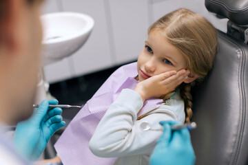 Child girl sitting in dental chair having pain in tooth, touching cheek and looking at male dentist