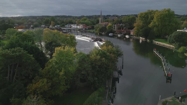 Autumnal trees frame a serene river scene with a weir and a charming town, including a church spire, under a cloudy sky.