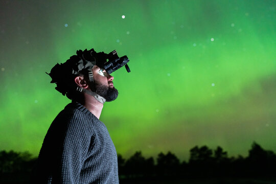 Side profile of a man using a professional night vision monocular device under a glowing green Aurora Borealis sky.