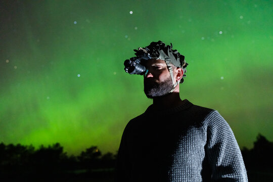 Close-up of a man in a textured sweater wearing a single-tube night vision device, looking up at a vibrant green Aurora Borealis night sky.