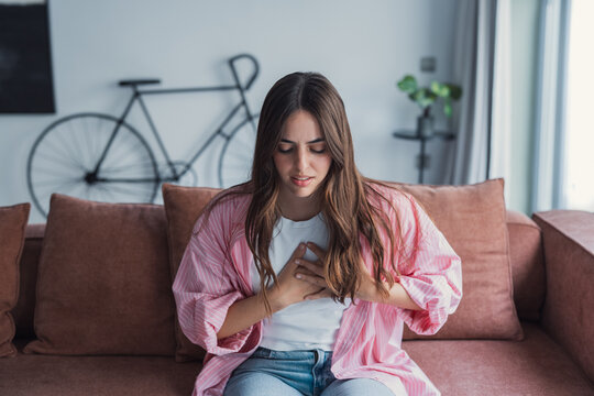 Young woman sits on a sofa clutching her chest and wincing, conveying chest pain, anxiety and possible heart-related discomfort in a casual home setting for health, medical and wellness topics.