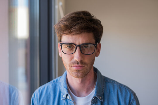 Thoughtful young man wearing glasses and a denim shirt looks toward the camera by a window, conveying introspection, focus and quiet confidence in a natural indoor portrait.