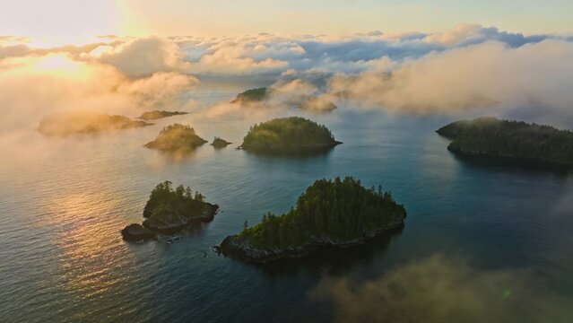 Aerial view of fog banks parting over calm ocean revealing lush floating archipelagos in golden sunrise light