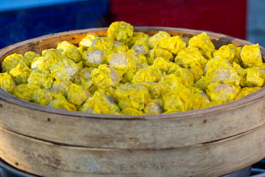 Freshly steamed yellow Siu Mai dumplings, a popular street food, are ready to be served from a traditional bamboo steamer basket at a vibrant Thai food market.