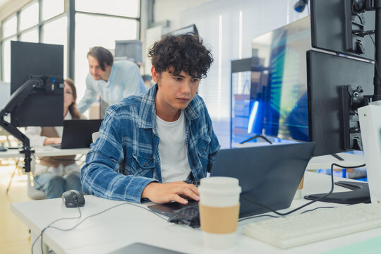 focused young male developer with curly hair in blue plaid shirt intensely codes on laptop in modern AI office, surrounded by multiple monitors displaying code neural networks and data graphs