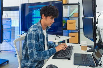 Fototapeta premium focused young male developer with curly hair in blue plaid shirt intensely codes on laptop in modern AI office, surrounded by multiple monitors displaying code neural networks and data graphs