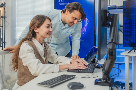 female developer in vest codes on laptop while male colleague in blue shirt leans over pointing at AI data visualizations on multiple monitors in modern tech office with large AI Assistant screen