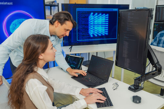 female developer in vest codes on laptop while male colleague in blue shirt leans over pointing at AI data visualizations on multiple monitors in modern tech office with large AI Assistant screen