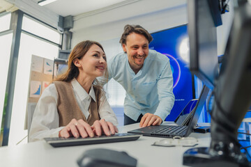 Fototapeta premium female developer in vest codes on laptop while male colleague in blue shirt leans over pointing at AI data visualizations on multiple monitors in modern tech office with large AI Assistant screen