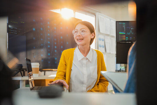 collaborative female AI developer in yellow sweater and glasses discusses code and neural network diagrams on large monitor while taking notes on tablet in modern tech office, with team members listen