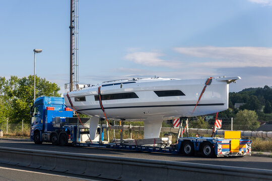 Special heavy truck transports sailing yacht hull on low trailer along highway through countryside. Secured yacht hull moves as convoy with escort along motorway under clear sky during transport