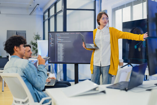 confident female AI team leader in yellow cardigan and glasses presents neural network architecture and code on large monitor while explaining to seated developers with laptops and tablets in office