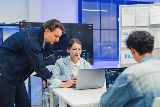 collaborative AI development team in modern tech office: young male developer coding on laptop, female team leader in light blue blazer pointing at code and neural network visualization 