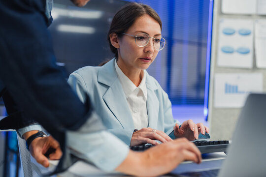 collaborative AI development team in modern tech office: young male developer coding on laptop, female team leader in light blue blazer pointing at code and neural network visualization 
