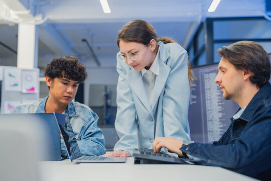 collaborative AI development team in modern tech office: young male developer coding on laptop, female team leader in light blue blazer pointing at code and neural network visualization 
