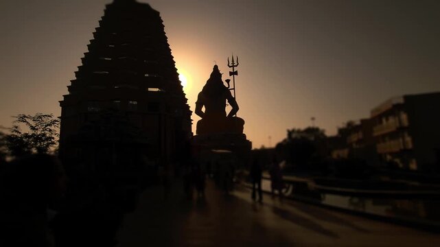 Silhouette of Lord Shiva statue with trishul near a temple during golden sunset in India. Spiritual Hindu scene with devotees, dramatic sky, Chaar Dham Temple in Vrindavan, Uttar Pradesh.