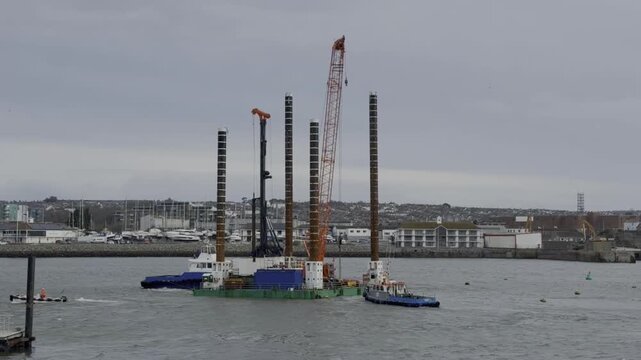Two tugboats pushing and pulling a large jack up rig, a specialized construction barge with tall spud legs and a crane, maneuvering it for positioning in the harbor for marine engineering work