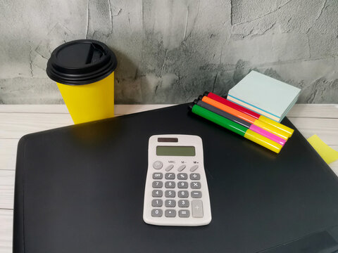 White calculator with markers, sticky notes and yellow coffee cup on black desk. Gray textured wall. Office supplies for work, study, business, finance, accounting