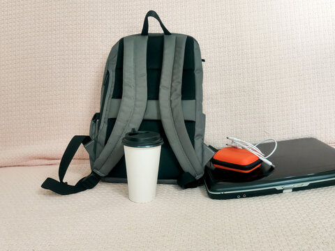 Gray backpack with coffee cup and black laptop with orange case on beige textured surface. Student essentials for school, university, remote work, travel. Mobile workspace for study tasks.