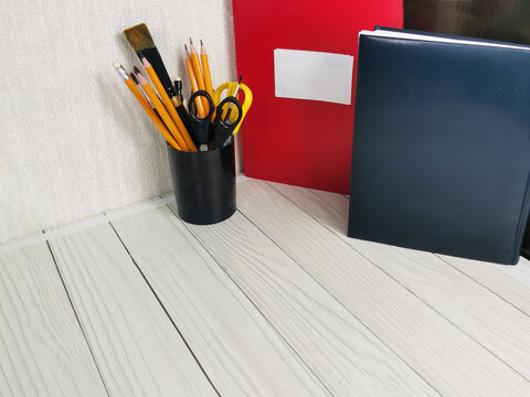 notebooks, pencils, scissors and paint brushes in black holder on white wooden desk. Office supplies for work, study, business organization, education workspace setup