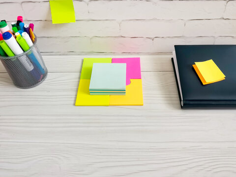 black notebook with sticky notes and colorful markers in holder on white wooden desk. Brick wall background. Office supplies for work, study, business planning