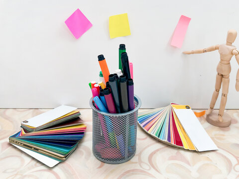 Colorful markers in mesh holder with color swatches and wooden mannequin on desk. Sticky notes on white wall. Design, creative work, art projects, color selection workspace