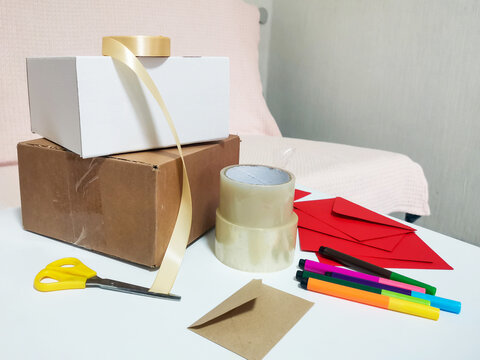 White and brown boxes with gold ribbon, tape, yellow scissors, red envelopes and colorful markers on white desk. Gift wrapping, crafting, DIY projects, creative workspace supplies