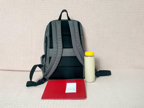 Gray backpack with red folder for documents and water bottle with yellow cap on sofa. School supplies, study accessories, student essentials, education, work commute, travel preparation