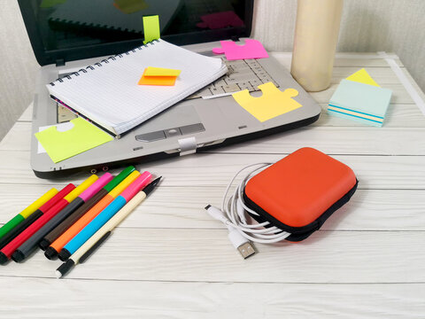 Gray laptop with colorful sticky notes, spiral notebook, markers and orange cable case with USB cords on white wooden desk. Remote work, study, education workspace setup