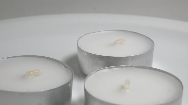 Close-up of three white tea light candles in silver aluminum cups on a white plate, captured on a rotating surface against a neutral grey background.