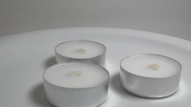 Close-up of three white tea light candles in silver aluminum cups on a white plate, captured on a rotating surface against a neutral grey background.