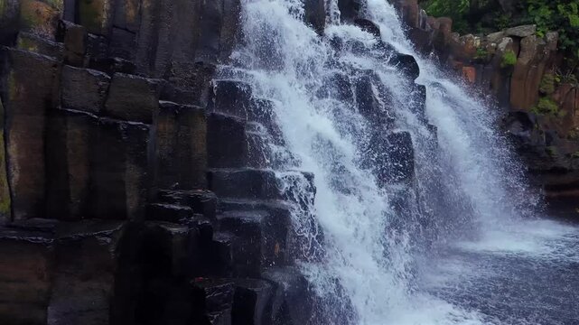 Water cascades down rugged basalt columns. Moss clings to dark, weathered rock faces. Water splashes and churns at the base. Scene exudes wild, untamed nature