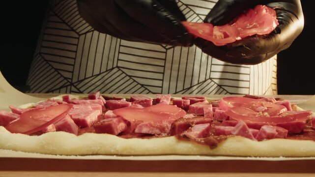 Lowangle closeup of layering cured meat in meticulous rows on pizza dough. Hands of chef in patterned apron and black gloves place tomato slices on top in kitchen. Arranging ingredients before baking