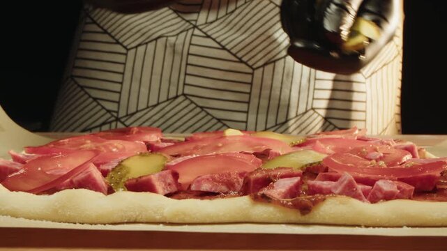 Closeup of hands in black gloves that add pickle slices on raw pizza dough with thin cured meat, tomato on wooden board. A chef in an apron stands at the table and prepares a traditional Italian dish