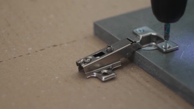 Close up of installing a furniture hinge on a cabinet door using an electric screwdriver. Process of screwing a self-tapping screw into hardware during furniture assembly.