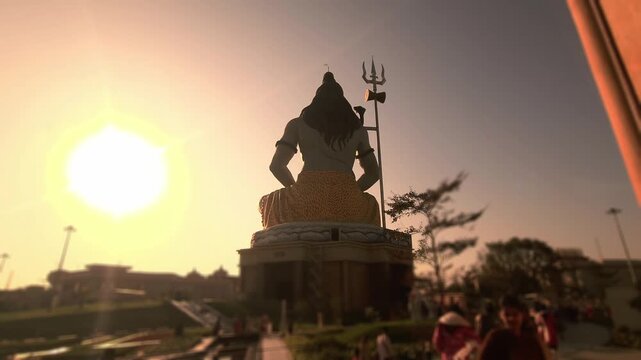 Silhouette of Lord Shiva statue with trishul near a temple during golden sunset in India. Spiritual Hindu scene with devotees, dramatic sky, Chaar Dham Temple in Vrindavan, Uttar Pradesh.