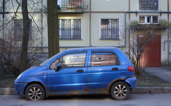 An old blue rusty car is parked near a residential building, Novocherkassky Avenue, Saint Petersburg, Russia, March 22, 2026