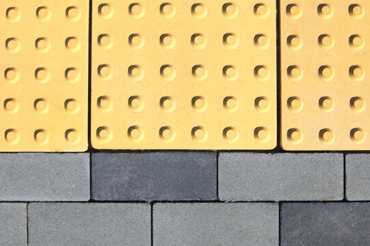 Yellow concrete paving slabs with convexed points. Special tactile paving slab with ribbed surface for blind people. Cover of grey rectangular paving slabs with embedded tactile concrete slabs
