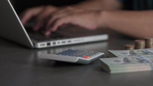 A hand pressing buttons on a calculator and calculating or typing transactions into a computer, with a pile of coins and banknotes beside it. Concepts related to banking and finance.